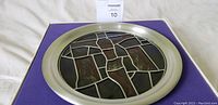 Circular stained glass plate with metallic frame on purple fabric background inside original box.