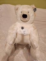 White plush teddy bear sitting upright on a beige bedspread, showing full front view with tags attached to ear and chest.