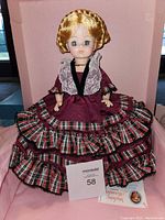 Front view of Madame Alexander Harriet Lane doll dressed in burgundy and plaid historic style gown with lace accents, sitting in pink box.