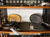 Cast iron pans including skillets, an oval pan, and a wooden tray in the background on a shelf