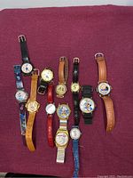 Top view photo showing all 12 Disney character wristwatches with varying bands and face styles placed on a maroon fabric background.