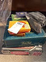 Multiple board and card games stacked including Sudoku, Trivial Pursuit game box, card decks, and mesh bag containing cards or pins.