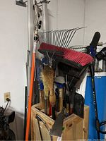 View of garden tools including brooms, rakes, pruning shear, shovel inside wooden storage crate in garage setting.