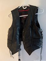 Front view of black leather Milwaukee motorcycle vest on hanger, showing snap button closures and lace-up sides.