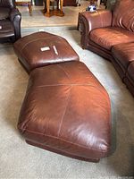 Two matching brown leather ottomans shown side-by-side on carpeted floor near leather furniture. Both have visible wear and need cleaning.