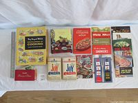 Top view of all 16 cookbooks and booklets arranged in one row showing covers and variety of cooking topics.