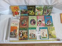 Front view of 15 books arranged horizontally showing titles including The Bobbsey Twins series, Little Women, Good Wives, Cherry Ames, and others, placed on a white backdrop with a ruler for scale.