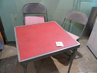 Photo showing vintage child-sized square card table with red vinyl surface and gray metal legs, accompanied by 2 folding chairs with red and white checkered padded seats and gray metal frames.