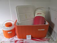 Four vintage Coleman cooler products including large orange cooler, orange jug, red jug, and umbrella.