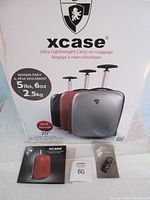 Photo shows original packaging box of Heys xcase carry-on luggage with three color options displayed and luggage specs including weight 5 lbs 6 oz, and size 20"x14"x9". Several original paperwork items and combination lock are placed in front.