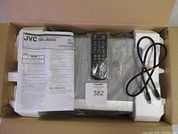 Photo showing boxed JVC HR-J693U VHS recorder with instruction manual, remote controller wrapped in plastic, and AV cable secured with rubber band inside packaging foam.