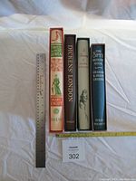 Four Folio Society classic books vertically aligned with rulers for scale and identification. Titles visible: Travels in West Africa, Dickens' London, Faust, The Spy's Bedside Book.