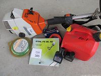 STIHL FS36 orange and white gas trimmer laying on surface with extra line, 2-stroke oil, red gas can marked 50:1, and manual.