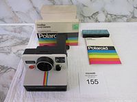Front view of the Polaroid OneStep Land Camera with the box behind it, instruction manual and unused flash unit beside it.