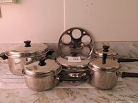 Set of stainless steel pots, frying pan, lids, and Androck Canada utensils with holder arranged on countertop.