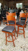 Three wood framed bar height chairs with black vinyl upholstered seats and some scratches visible on the wood frames. Chairs arranged outdoors on gravel surface with workshop background.