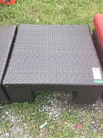 Square garden table made of faux rattan material, placed outdoors on grass and gravel.