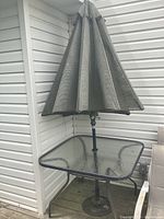 Image showing square glass topped patio table with umbrella fully closed, placed on a wooden deck corner beside house siding and outdoor chairs