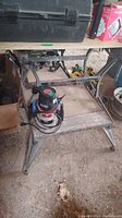 Photo showing Black and Decker Workmate work table with Sears Craftsman router placed on bottom shelf, visible cords, metal frame with wooden surface.