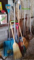 Full view of various garden tools including brooms, shovels, and rakes leaning against a wall