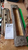 Photo showing two pieces of vintage metal door hardware including a large Victorian house number frame and a smaller keyhole plate on a wooden surface.