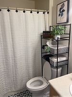 Bathroom corner showing black metal shelf with storage containers, faux books, and green plant, white textured shower curtain, floor tile, and framed botanical art on the wall.