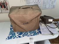 Sewing machine inside beige fabric cover placed on blue and white floral cloth on table, with power pedal and documents nearby.