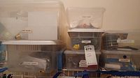 Wide view of multiple clear plastic storage containers stacked on a shelf, each labeled with contents such as light bulbs, sticky stuff (adhesives), cords, nails, screws, washers, and gliders (furniture pads).