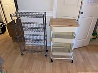 Photo showing two kitchen storage carts on casters side by side on wooden floor against white wall and door.