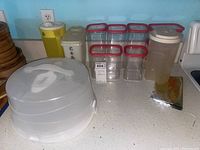 Image showing multiple clear plastic containers with red lids, two opaque storage containers with white lids, a white and yellow pitcher, and a multi-tiered plastic cake carrier with cover on a kitchen counter.