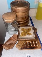 Glass pitcher with cork lid, set of four bamboo steamers, multiple wooden cutting boards, and ceramic trivet with leaf design arranged on a kitchen counter.
