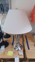 Two metal table lamps side by side on a wooden surface. Both have white fabric shades and brushed metal bases, cords are visible but wrapped around the bases.