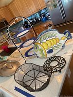 Decorative fish-shaped ceramic plate with lemon and leaf design next to a three-tier metal plate stand with blue and white plates, a round black metal trivet with scroll design, and an oval black wire basket holder on a kitchen counter.