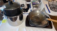 Overview image showing stacked stainless steel pot, glass lid, Helen Chen wok with wooden handle on top along with several baking sheets underneath.