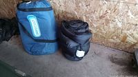 Photo of a black lightweight sleeping bag rolled and secured, placed beside a blue Aero air bed carry bag.