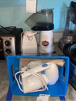 Photo of Cuisinart hand mixer, electronic can opener, and air popcorn maker stacked on countertop with blue basket containing hand mixer and beaters visible.