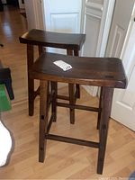 Front and angled view of two wooden stools with curved seats and dark finish. Shows pattern of wear and scratches on surfaces and legs.