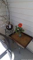Photo showing black metal plant stand with brown vase and faux branches, small square table with wooden tray and green vase with faux orange flower