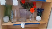 Seven vases on a wooden shelf featuring varying colors and styles including green matte, clear glass, white ceramic, and blue-green artistic glass.