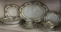 Porcelain dish set with plates, cups, saucers, oval serving platter, covered bowl, and serving bowls displayed on shelf showing floral and gold trim detail.