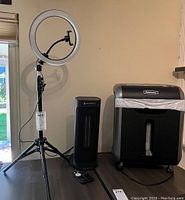 Full view of the three main items in the lot: adjustable ring light on tripod, black space heater with remote, and Fellowes paper shredder with waste bin and wheels.