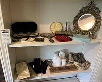 Overview photo of lot items displayed on shelf including dresser mirror, jars, gloves, baby shoes, grooming tools, cribbage set