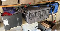 Garage shelf with gray trunk storage caddy, black bin containing assorted spray cans and bottles, and blue/black soft sided cooler bag on the right.