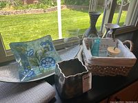 Photo showing curved blue floral glass dish, basket, vases, and placemats on countertop near window.