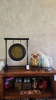 Wide view of top shelf showing metal gong with black stand, ceramic owl figurine, white pitcher, small container, coasters, and small figurines placed on wooden furniture.