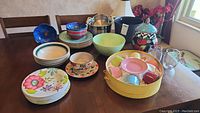 Photo showing assortment of ceramic, stoneware, and plastic dishware including plates, bowls, stock pot, cookie jar, coffee cup set, and drinking glasses on wooden table
