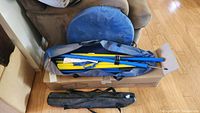 Several poles in yellow and blue colors inside a blue carrying bag, alongside a blue collapsible tunnel lying next to a chair on wooden floor.