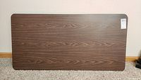 Rectangular metal folding table top with dark brown wood grain finish, placed on carpeted floor.