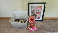 Plastic tote with assorted small picture frames, large framed print with cat and floral design, pink metal flower wall hanging, and floral fabric piece arranged on carpeted floor.