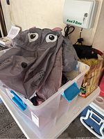 Folded gray inflatable hippo costume with black and white eyes, stored in a clear plastic bin with blue handles, with some household background visible.